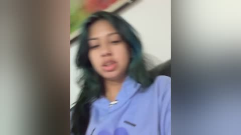 Video of an Asian woman with long, wavy, green hair, wearing a light blue shirt, standing in a hallway with blurred, colorful artwork on the wall.