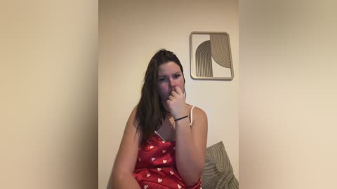 Video of a young woman with long brown hair, wearing a red tank top with white polka dots, sitting on a bed. She covers her mouth with her right hand, wearing a bracelet on her left wrist. Background features a beige wall with a geometric art piece.
