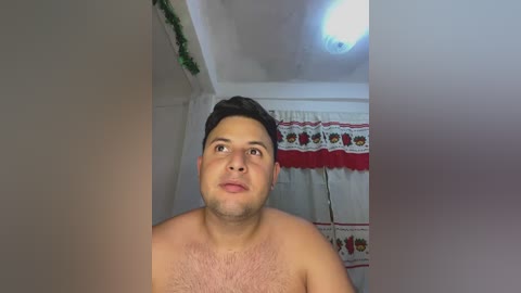 Video of a shirtless, light-skinned man with short dark hair, looking up with a neutral expression. Background features a white curtain with red and green floral pattern and a small, bright light fixture.