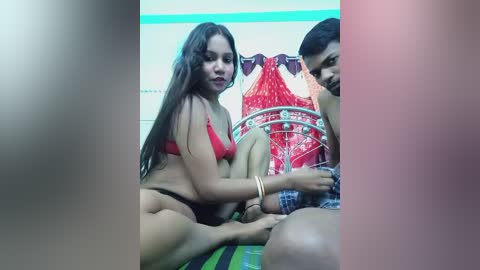 Video of a young South Asian woman in red lingerie, sitting on a bed, with a man's bare legs visible in the foreground.