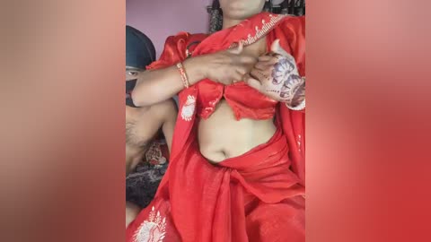 Media: Video of a woman in a red sari, partially lifting it to reveal a tattoo on her midriff. She has a fair complexion and wears a bracelet. Background shows a pink wall and a dark object.