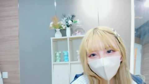 A video of a young woman with long blonde hair, wearing a white mask, standing in a modern, minimalist bathroom with grey walls, white shelving unit, and decorative flowers.