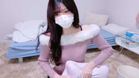 Video of an East Asian woman with long dark hair, wearing a white face mask, pastel pink sweater with white fur trim, and white pants, kneeling on a beige carpet in a minimalist bedroom with a bed, nightstand, and a glass table.