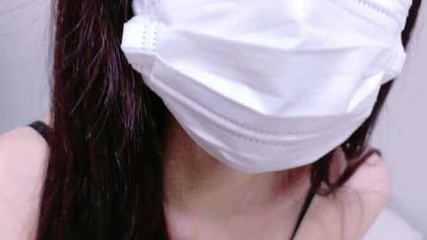 A close-up video of a person with long, wet, dark brown hair, wearing a white surgical mask, and a black spaghetti strap top, with visible skin and a slight redness on the neck.