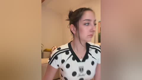 A video of a young woman with auburn hair in a ponytail, wearing a black and white polka-dotted shirt, standing in a dimly lit room with beige walls.