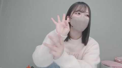 A video of an East Asian woman with straight black hair, wearing a white mask and a pink sweater, making a peace sign with her right hand, against a plain, light gray background.