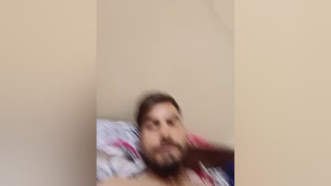 A blurred video shows a man with a beard lying on a bed, partially covered by a white and red patterned blanket, against a beige wall.