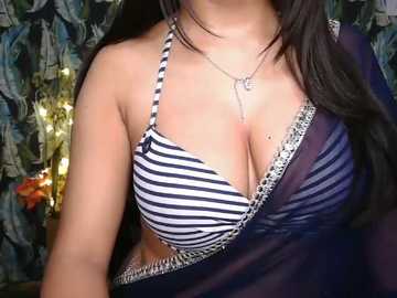 Video of a woman with medium skin tone wearing a black and white striped bra with silver chain detailing. She has long, dark hair, and wears a delicate necklace. The background features floral-patterned curtains and a warm, ambient light.