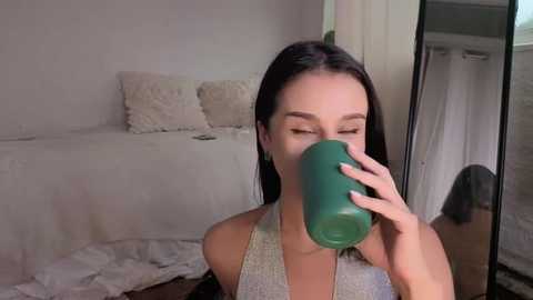 A video shows a young woman with straight dark hair, wearing a silver halter top, drinking from a green cup, in a minimalist, softly lit bedroom with white bedding and a mirror.