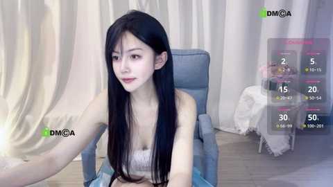 Video of a slender, fair-skinned woman with long black hair, wearing a white lace bra, seated in a blue chair, against a soft-focus background.