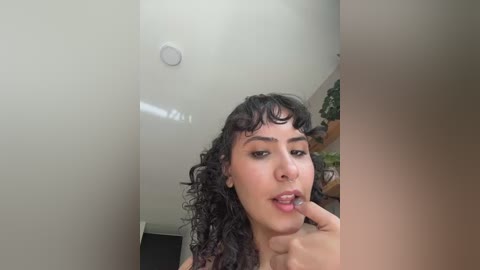Media: A video of a young woman with curly dark hair and light skin, wearing a nose ring, in a room with white walls and a potted plant.