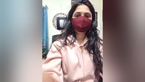 Video of a young woman with long black hair, wearing a maroon face mask and pink hoodie, standing in a room with a blue curtain and fan.