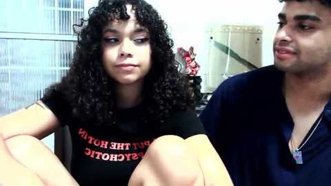 Video of a young Latina woman with curly black hair and light skin, wearing a black T-shirt, sitting next to a young Latino man with short black hair and a navy shirt, in an office setting.