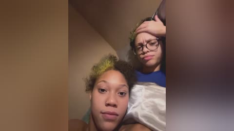 Video of two young African American girls with curly hair; one with green hair, the other with glasses, both wearing casual clothes, in a dimly lit room.
