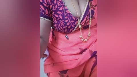 Video of a woman wearing a vibrant, low-cut, floral-patterned blouse and a flowing, coral saree. She has a gold necklace with tassels, and the background is a gradient of pink hues.