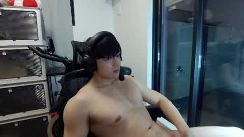 Video of a nude, muscular Asian man with medium build and small breasts, wearing black headphones, sitting on a gaming chair in a modern, minimalist room with white walls and black furniture.