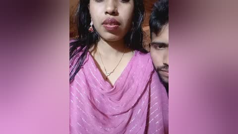 Video of an Indian woman with long black hair, wearing a pink, sheer sari with subtle white patterns. She has a sad expression and a red lipstick. Beside her, a man with a mustache looks concerned.