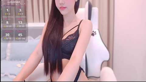 Video of an East Asian woman with long, straight black hair, wearing black lace lingerie, sitting on a white bed in a softly lit, modern bedroom.