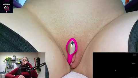 Video of a close-up view of a woman's shaved vulva with a pink anal plug inserted. The image includes a video feed of her from a webcam.