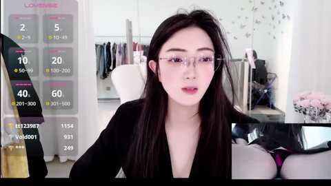 Video of a fair-skinned, long-haired Asian woman wearing glasses, a black top, and pink underwear, indoors with a calendar, clothing rack, and flowers in the background.