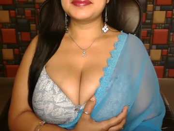 Video of a South Asian woman with long black hair, wearing a blue sheer blouse over a gray lace bra, showcasing ample cleavage. Background features a colorful mosaic wall.