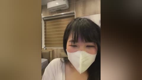 A video of an Asian woman with long black hair, wearing a white mask and white shirt, sitting in a beige-colored room with wooden paneling and an air conditioner.