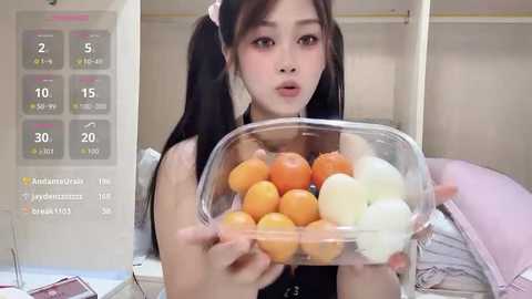 A video of a young Asian woman with long black hair in pigtails, wearing a black top, holding a clear plastic container filled with colorful fruit (oranges, apples, and bananas) in a hospital room.