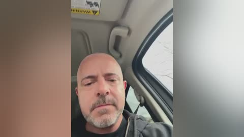 Video of a bald, middle-aged Caucasian man with a mustache and glasses, wearing a black shirt, sitting in a car, eyes closed, possibly sleeping. The background shows a beige car interior.