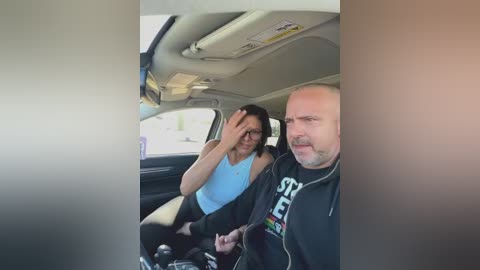 Video of a man and woman sitting in a car; he's bald with a beard, wearing a black hoodie, and she's wearing glasses and a blue tank top.
