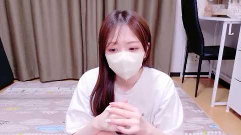 Video of a young Asian woman with long brown hair, wearing a white mask, sitting on a gray-patterned rug, indoors with beige curtains and a black chair in the background.