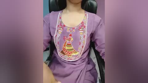 Video of a person in a purple dress with intricate embroidery featuring a red and yellow dancer, seated in a black chair, with blurred faces in the foreground.