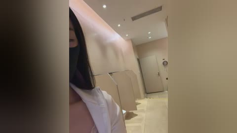 Video of a woman with long black hair partially covered by a white towel, standing in a beige-tiled bathroom with recessed lighting and a mirror.
