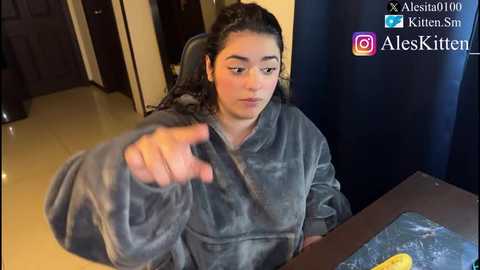 Video of a young woman with curly hair, wearing a grey fuzzy robe, gesturing with her right hand. She's seated at a dark wooden table in a dimly lit room with cream-colored walls.