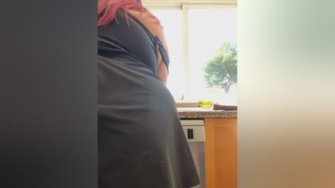 A video of a person with red hair wearing a black apron, bending over a kitchen sink with a window behind, showcasing a bright, sunny day with greenery outside.