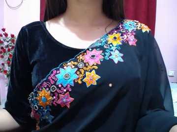 Video of a woman with long dark hair, wearing a black velvet blouse adorned with colorful embroidered star motifs in pink, yellow, and blue. Background features a red curtain and a floral arrangement.