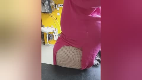 Video of a person wearing a pink dress with beige mesh paneling over their buttocks, taken indoors with a yellow wall, sink, and clothes hanging in the background.