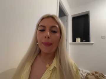 A video of a blonde woman with long hair, fair skin, and subtle makeup, wearing a yellow blouse, smiling in a simple, dimly lit room with a window and a light switch visible.