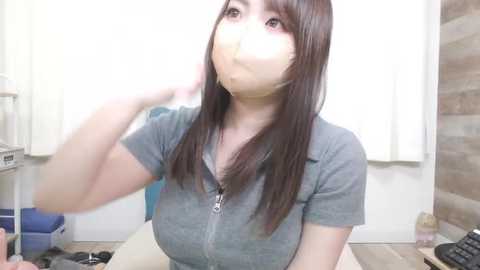 Video of an Asian woman with long brown hair, wearing a gray shirt, with a large piece of tape over her mouth, seated indoors with a white wall and brown wooden panel background.