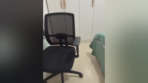 Video of a minimalist, modern bedroom with a black mesh office chair and a neatly made bed with a light green comforter. The background features white wardrobes and a beige tiled floor.