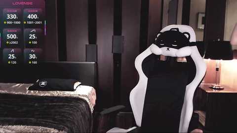Video of a plush panda bear in a black gaming chair in a dimly-lit bedroom with a bed, nightstand, and digital display.