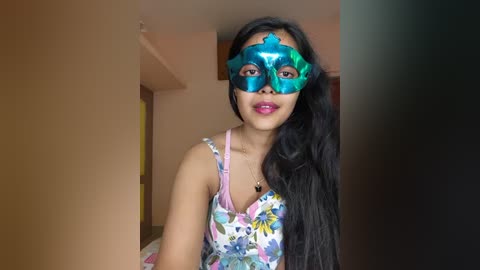 Video of a young woman with long black hair, wearing a blue Venetian masquerade mask and a colorful floral top, standing in a simple, beige-walled room.