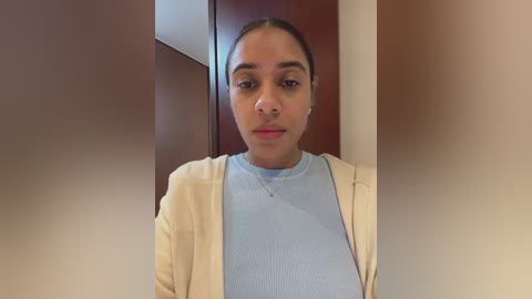 Video of a young woman with medium brown skin, dark hair pulled back, wearing a light blue ribbed sweater and beige cardigan, standing indoors against a blurred background.