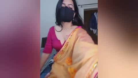 A video of a South Asian woman with medium skin tone, black hair, and dark eyes, wearing a red blouse, black face mask, and a yellow sari with colorful embroidery, in a dimly lit room with a partially open door and a hanging garment.