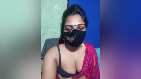 Video of a South Asian woman with medium-dark skin, wearing a black mask, red saree, and a black bra, sitting against a green and blue wall.