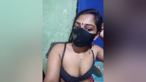Video of a South Asian woman with medium-dark skin tone, wearing a black mask, black bra, and a necklace, in a modest, dimly lit room with green and blue walls.