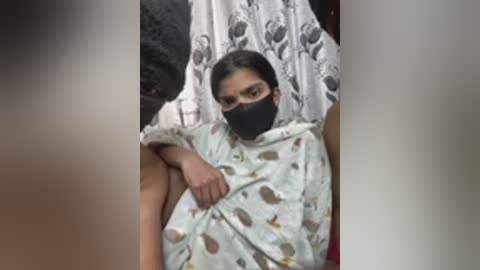 Video of a woman with dark skin, wearing a black face mask and a light-colored patterned dress, sitting on a white chair with a floral patterned backdrop.