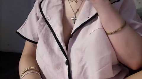 A close-up video of a light-skinned woman in a light pink blouse with black trim, wearing a cross necklace and a bracelet.