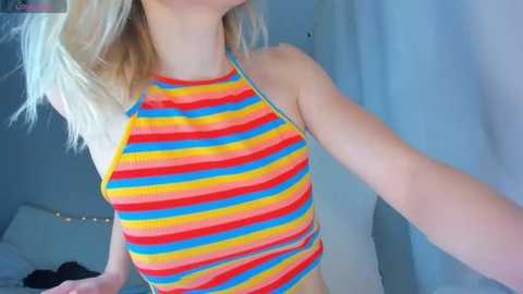Video of a young woman with fair skin, wearing a colorful, striped halterneck swimsuit, standing indoors with a white curtain in the background.