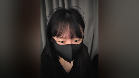 A video of an East Asian woman with long black hair, wearing a black mask and dark clothing, gazing intently, framed by gray curtains in a dimly lit room.