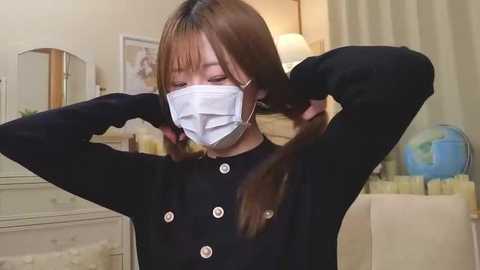 Video of an East Asian woman with straight brown hair, wearing a black cardigan, white face mask, and gloves, adjusting her hair in a cozy, softly lit bedroom with cream-colored furniture and a globe.